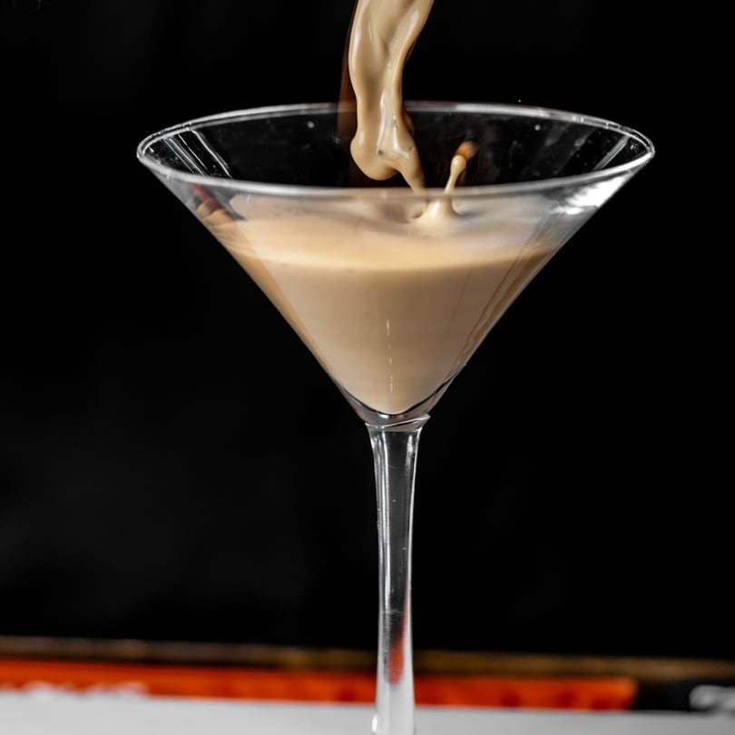 Creamy beige liquid pours into a martini glass, creating a splash against a dark background.
