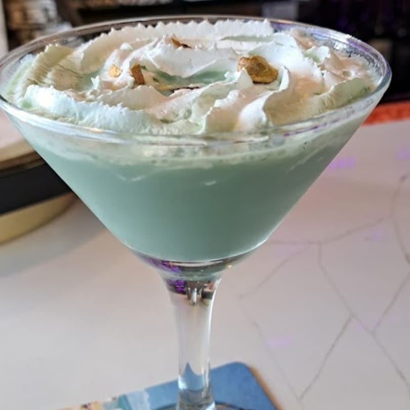 A light green martini with whipped cream and gold sprinkles sits on a white bar.