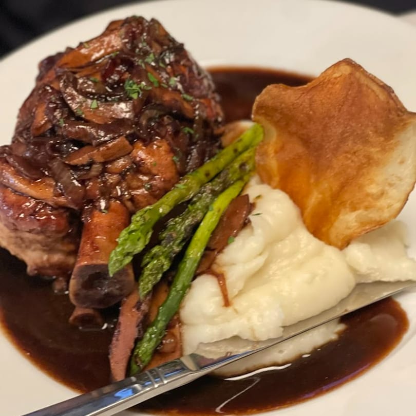 Braised lamb shank with mushrooms, asparagus, mashed potatoes, and a crispy potato chip on a white plate.