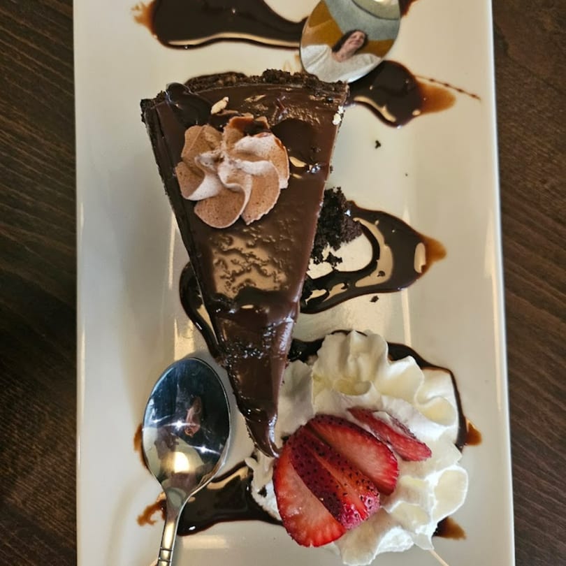 A slice of rich chocolate cake with whipped cream, chocolate drizzle, and strawberries on a white plate.