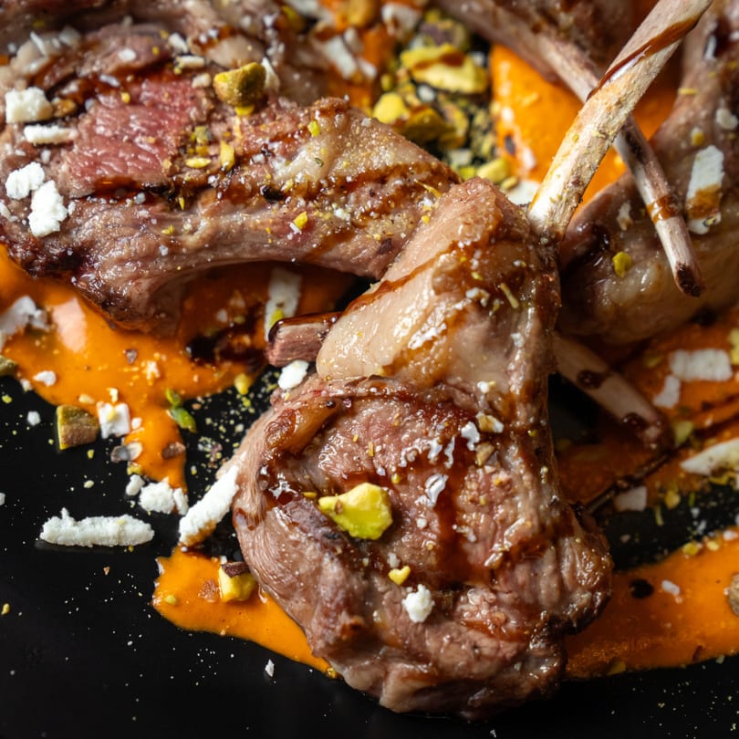 Grilled lamb chops with orange sauce, crumbled cheese, and chopped pistachios on a black plate.