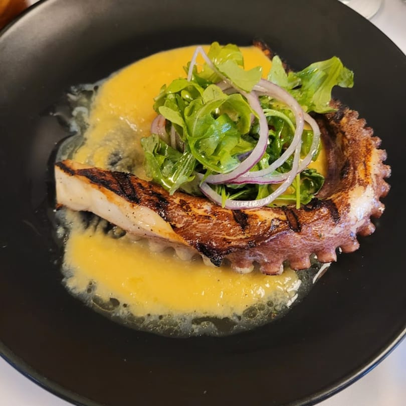 Grilled octopus with a vibrant yellow sauce, topped with fresh arugula and red onion slices.